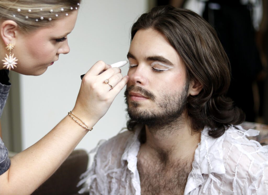 Photo of Jackson Riddle getting his eyemakeup done by a makeup artist.